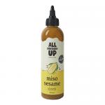 A clear glass bottle holds pale beige miso and sesame dressing speckled with black sesame grains. A black screw cap tops the bottle. The label shows the brand name in bold script, minimal icons, and clean typography with earthy beige and dark brown accents. Glossy highlights and sesame flecks shine.