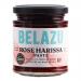 A neat arrangement of clear glass jars with black lids shows vibrant rose-red harissa paste. Each jar displays a Belazu label with simple typography and small floral detail. The glossy, textured paste fills the jars, catching light and revealing flecks of chilli and spices. On a soft cream backdrop.