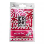 Bright red velvet snack bars sit in glossy wrappers arranged in two neat rows inside a rectangular box. Crumb-speckled surfaces peek through transparent windows. Packaging features cream accents and simple branding, with one unwrapped cube showing dense, moist texture. A small nutrition label shows.