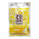 A stack of yellow Coco Cubes Banana wrappers rests on a light surface. Square coconut-and-banana pieces appear through small windows. Glossy packaging shows a brown logo, banana illustration and simple icons. Soft shadows and warm lighting create depth. Close-up shows coconut flakes and ripe banana.