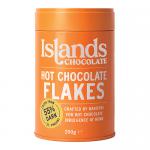 Photograph shows a 200g bag of Islands Chocolate 55% Dark Hot Chocolate Flakes. Transparent window reveals irregular dark brown flakes. A cream-and-brown label displays the brand name, '55% dark' text and an island graphic. The packet rests on a wooden surface with a few flakes scattered. Warm tones