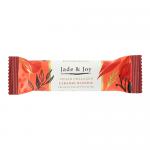 Wrapped bar leans against a stack of identical bars; glossy cream-and-teal wrapper features 'Jade & Joy' logo, caramel drip graphic and blondie image. Unwrapped bar reveals golden blondie with caramel swirls, crumbly texture and a light nutty topping. A minimal white backdrop and soft shadows accent