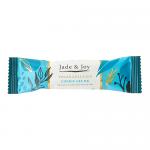 The photograph shows a stack of 12 Jade & Joy Vegan Collagen Cookie Crumb protein bars in glossy wrappers. Each wrapper displays a pale green logo, brown cookie crumb imagery and a beige bar illustration. One bar is partially unwrapped, revealing a crumbly, speckled interior Under soft studio light