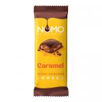 A large chocolate block sits in a warm-toned wrapper featuring the NOMO logo. The bar displays segmented squares with an embossed brand mark; glossy caramel ribbons thread between and across broken pieces, contrasting deep milk-chocolate brown with golden, sticky caramel highlights. Subtle dim sheen