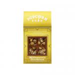 A clear bag of golden popcorn studded with crumbly shortbread pieces and glossy caramel and chocolate drizzle sits spilling onto a wooden surface. Bright branded packaging rests nearby with scattered crumbs and a paper scoop; warm light highlights texture and sheen. Soft reflections kiss the popcorn