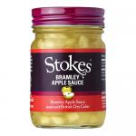 Six clear glass jars sit in a cardboard tray, each filled with pale golden Bramley apple sauce. Green-and-white labels wrap the jars with the Stokes name; silver lids catch the light. Jars stand closely together, revealing a smooth, slightly chunky fruit texture.