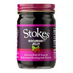 Six clear glass jars sit in a tidy row, each holding deep ruby-red jelly. Silver lids reflect light. White labels display the Stokes logo and simple black text. Light catches the glossy surface of the jelly, revealing smooth, translucent texture and tiny suspended seeds. Thin paper bands around base.
