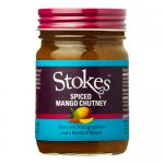 Clear glass jar shows chunky, golden-orange mango chutney. A cream-and-red label wraps the middle with the Stokes name and product text; a dark metal lid closes the top. Visible fruit pieces sit in glossy, spiced syrup and light reflects off the textured surface. Label typography and crest look crisp