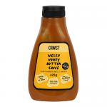 A clear glass jar with a metal screw lid displays a glossy amber honey-butter sauce. A cream label shows bold CRWST lettering, subtle Welsh emblems and handwritten-style script. Light catches the smooth, viscous surface and a dab clings to the jar rim. A soft shadow falls on a neutral pale backdrop.
