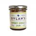 A clear glass jar holds glossy deep-red chilli jam studded with visible seeds and chili fragments. A white label wraps the jar with bold brand lettering and the product name. A metal screw lid tops the jar and casts a soft reflection.