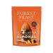 A glossy milk chocolate slab studded with whole almonds and vivid Valencia orange shards lies partly unwrapped beside a neat stack of Forest Feast Fruit, Nuts & Seeds packs; warm-toned, rustic artwork on the packaging depicts fruit, nut and seed illustrations.