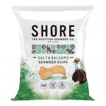A small matte pouch with a coastal logo lies open, spilling irregular glossy dark-green seaweed crisps dusted with coarse white salt and streaked with balsamic sheen. Crinkled edges, varied textures and scattered salt crystals contrast with the clean, seaside-inspired packaging.