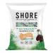 A small matte pouch with a coastal logo lies open, spilling irregular glossy dark-green seaweed crisps dusted with coarse white salt and streaked with balsamic sheen. Crinkled edges, varied textures and scattered salt crystals contrast with the clean, seaside-inspired packaging.