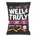 Well&Truly Crunchies Smokey Bacon packet lies on a pale surface. Red and brown packaging shows a prominent logo and product name, vivid photos of ridged crunchy strips, wisps of smoke graphics and scattered bacon pieces. A clear 100g weight mark sits in a corner. Soft highlights pick up slight sheen
