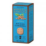 A neat row of six round, golden-brown biscuits sits beside a rustic bakery box labeled 'Artisan Biscuits'. Each biscuit shows a glossy spiced caramel glaze, specks of spice and sugar crystals, with crisp edges and a crumbly, buttery interior visible.