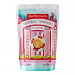 A glossy box and opened tray reveal golden round biscuits topped with crumbly crust and creamy pink swirls. The packaging features vintage bakery lettering, a strawberry cheesecake illustration with sliced berries, pastel pink accents and a clear bold product name across the front. Subtle rim light.