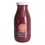 A cluster of clear glass jars sits on a neutral surface. Vivid rhubarb and beetroot ketchup shows as deep pink-red through the glass. Each jar wears a cream label with neat typography and a small coastal emblem. Light glances off the glossy sauce and metal lids. Sea-salt flecks dot the paper labels.