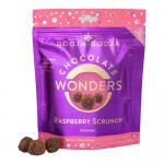 A photo shows a matte brown box of Booja-Booja Raspberry Scrunch Chocolate Wonders with soft pink branding. Eight dark chocolate rounds sit beside the open box; several are unwrapped to expose a crumbly raspberry-pink filling. Raspberries and crumbs dot a pale wooden surface.
