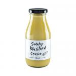 A clear glass jar with a metal lid contains smooth, amber-brown smoky mustard. A cream-and-black label encircles the jar, displaying the Hawkshead Relish name and stylized mustard motif. The sauce shows fine dark speckles and a glossy surface reflecting light. A ribbed, metal lid shows subtle wear.