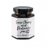 A glass jar of Hawkshead Relish Sour Cherry & Prosecco jam sits on a wooden board, its deep ruby preserve visible through clear glass. A white lid shows a neat label; scattered sour cherries and a small sprig of green leaves add natural color contrast. Soft light accents glossy texture, fine bubbles