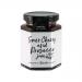 A glass jar of Hawkshead Relish Sour Cherry & Prosecco jam sits on a wooden board, its deep ruby preserve visible through clear glass. A white lid shows a neat label; scattered sour cherries and a small sprig of green leaves add natural color contrast. Soft light accents glossy texture, fine bubbles
