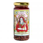 A glass bottle of crispy chilli oil sits against a neutral background. Deep red oil holds suspended flakes and seeds. A vintage-style label shows a stylized Mary Magdalene portrait framed by ornate lettering and subtle gold accents, capped with a black screw top.