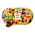 An open metal tin sits on a pale surface; its illustrated lid shows Parisian-inspired artwork and bold lettering. Ten dark chocolate truffles lie in a neat grid, glossy and dusted with cocoa, their deep brown contrasting with the colorful tin.