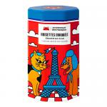 A round metal tin bearing Le Chocolat des Français logo and playful dog illustrations sits open, revealing glossy chocolate-coated hazelnuts. A few whole and halved nuts spill onto a light surface, showing rich brown coating and pale, textured interiors beside the colorful lid.
