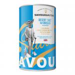 Photograph shows a cylindrical Savoursmiths tin of Desert Salt & Vinegar crisps with a matte pale-blue label featuring stylized desert dunes and salt crystals. Festive holly and subtle snowflakes frame the logo. Several tins sit stacked against a soft, neutral background. Bright twinkle lights glow.
