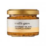 A set of six 90g glass jars of Truffle Guys Gourmet Truffle Honey sits arranged in a row inside a festive gift box. Warm amber honey shows dark truffle flecks. Each jar bears a minimalist black-and-white label with the Truffle Guys logo and gold lids reflecting light. Soft highlights on glass gleam.