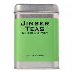 A festive box of Jinger Drinks ginger and mint tea sits on a table, bright red packaging with gold snowflakes and green mint illustrations. A steaming cup, sliced ginger and fresh mint leaves surround a neat row of tea bags beside the box Accents include twinkling lights and a single cinnamon stick