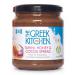 A glass jar sits on a plain background, showing a creamy, swirled spread in warm beige and chocolate brown tones. The label displays a rustic logo and product name in clean type. A wooden spoon rests beside the jar, coated in glossy, velvety spread, soft ripples and tiny cocoa flecks catching light.