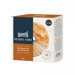 A sturdy rectangular box displays stacked round sourdough crispbreads with central holes. The crispbreads show a golden-brown, blistered surface, irregular air pockets and thin, crunchy edges. Minimal, rustic packaging and clear window reveal the textured rounds inside. Charred edges, small air holes