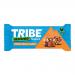 A single wrapped TRIBE x Wildfarmed flapjack sits on crumpled parchment beside an unwrapped oat bar studded with pecan halves and glossy raisins. Golden toffee glazes the surface, crumbs scatter, and bold TRIBE logo and flavour text appear on warm brown and cream packaging. Subtle gloss, oat crumbs.