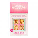 A photograph shows several Popcorn Shed Pink Gin packets with glossy pink and white wrappers decorated with holly and snowflake motifs. Loose pale-pink popcorn spills from an opened pack onto a wooden table, sprinkled with glittering sugar crystals and warm fairy lights. Soft-focus bokeh glows near.