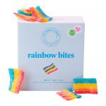 A glossy rectangular box displays a rainbow-striped background and a clear window revealing bite-sized round candies in assorted pastel colours. Playful cartoon characters and a bold white logo occupy the upper panel; ingredient and nutritional text appear on the rear, with small icons and barcode.