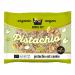 A round pistachio oat cookie rests on white parchment, showing a golden-brown surface studded with crushed green pistachio pieces and visible oat flakes. Crumbs scatter nearby; soft texture and rustic, home-baked appearance catch the eye. A Kookie Cat label peeks, minty accents mirror the pistachio.