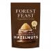 Bright matte wrapper shows stylized firs and deep-green forest graphics with a warm brown band and subtle logo. A milk-chocolate praline bar with whole hazelnuts sits front and center, one segment broken to reveal smooth, nut-speckled filling; six bars line up behind.