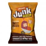 A bright orange bag of organic sweet potato crisps sits next to a small pile of thin, wavy, rust-colored chips. The crisps display ridges and visible salt crystals; a few whole sweet potato slices and scattered crumbs rest on a pale wooden surface under soft natural light. A few salt flakes glisten.