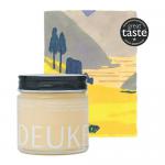 Six glass jars of Deukes Raw Monofloral Rapeseed Honey sit clustered on a wooden surface. Pale creamy-yellow honey fills each jar, showing smooth, opaque texture. White labels with simple black text and yellow accents wrap the jars; metal gold lids catch soft, natural light. Shallow blur behind jars