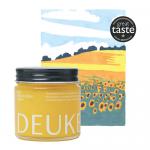 Photo shows six glass jars of Deukes Raw Monofloral Sunflower Honey arranged on a wooden surface. Golden honey gleams through clear glass, each jar bears a cream-and-yellow label and a metal lid, soft light highlighting the viscous texture.