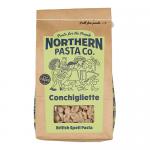 A clear plastic bag of small shell-shaped pasta shows tightly packed pale yellow conchigliette inside. A simple white label with blue and green accents names the brand and product, while nutrition facts and barcode appear on the back. The package sits on a plain surface. Soft shadows fall to the right.