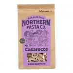 A stack of eight Northern Pasta Co. 450g packages fills the frame. Each crisply printed bag features the brand name, a clear window revealing short, twisted casarecce noodles in warm golden tones, and simple graphic accents. Lighting casts soft highlights on the textured pasta. Subtle shadows deepen.