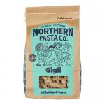 An image shows a clear bag of gigli pasta, revealing short, fluted trumpet-shaped pieces. A white label bears a modest logo and blue accents. The package sits on a neutral surface, warm light highlighting the golden-yellow ridged pasta.