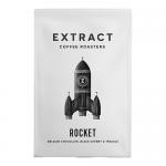 A sealed matte-black bag of Extract Coffee Roasters Rocket Espresso beans stands upright on a wooden shelf. A minimalist label shows a rocket logo and bold white text; a small pile of glossy dark-brown beans spills beside the bag.
