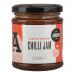 A clear glass jar of bright orange-red scotch bonnet chilli jam sits against a neutral background. The jam shows a chunky texture with visible seeds and pepper pieces. A bold label wraps the jar with brand lettering and a colorful chilli illustration beneath a black screw lid. Glossy surface gleams.