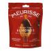 A glossy box labeled Meurisse sits beside a small pile of dark chocolate-covered almonds, each almond gleaming under light. The packaging uses deep browns and gold accents, with a clear window showing the confections.