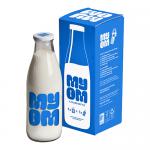 A clear 1000ml Myom glass milk bottle stands upright. It features a cylindrical body that tapers to a narrower neck with a defined rim, smooth reflective glass and subtle embossed branding. Soft highlights and shadows emphasize its contours.