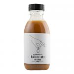 Several slender glass bottles sit upright on a wooden shelf, each filled with glossy dark brown sauce. Clean cream labels feature bold black lettering reading Peckham Sauce Co. with a small red accent. Matte black caps and a subtle hand-drawn illustration complete the tidy, artisanal look. Warm hue.