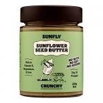 Six clear jars of Sunfly Sunflower Seed Butter sit in a row, each jar revealing a light-brown, crunchy spread flecked with sunflower seed pieces. Bright yellow sunflower graphics and bold Sunfly branding wrap the labels; metallic lids catch soft studio light. Soft reflections highlight jar contours